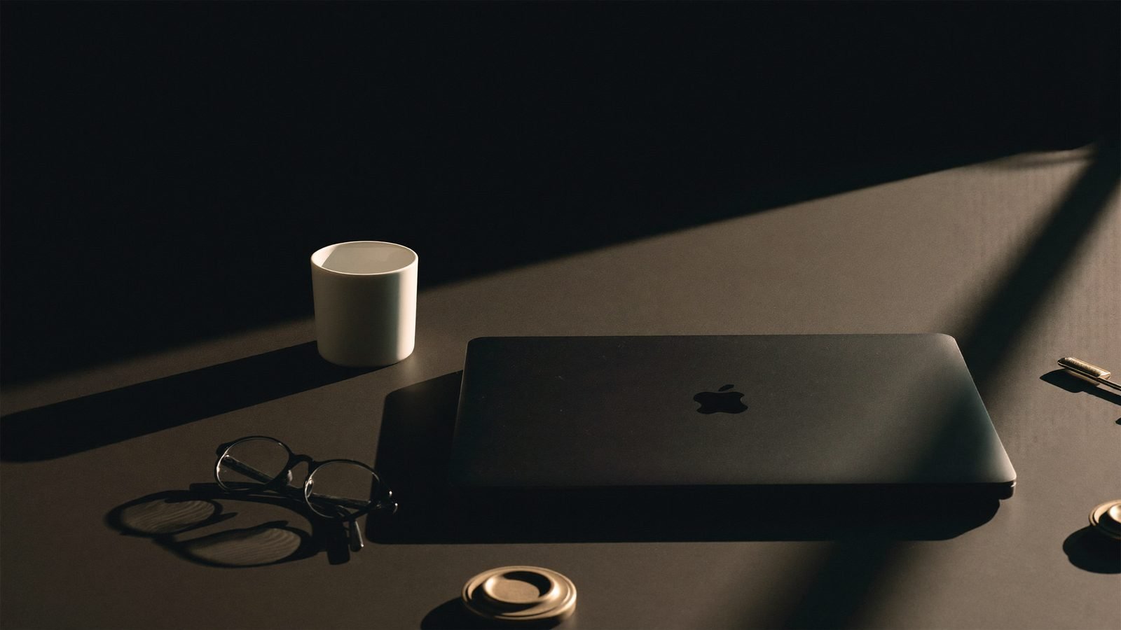 A closed laptop, reading glasses and a cup on a dark desk lit by a single shaft of window light